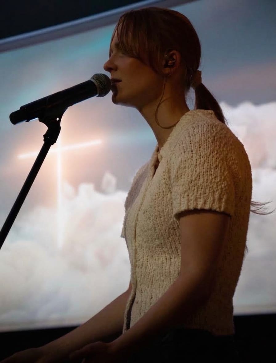 Ema in profile at a microphone stand, a cross projected on the screen behind her